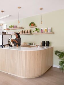 Barista at the curved fluted wood counter, with shelves of coffee and plants against the green wall