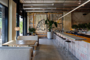 Bar at MAMENORI with a tiger artwork on a tiled mosaic wall, an aluminium profile bar top, a terracotta-coloured block bar front and steel stools under suspended linear lights